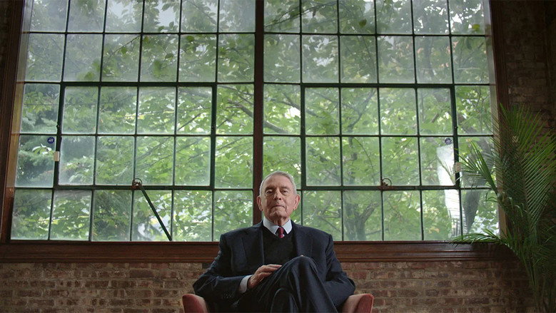 Dan Rather in a modern newsroom, his posture slightly hunched but his expression still sharp, surrounded by the glow of television monitors