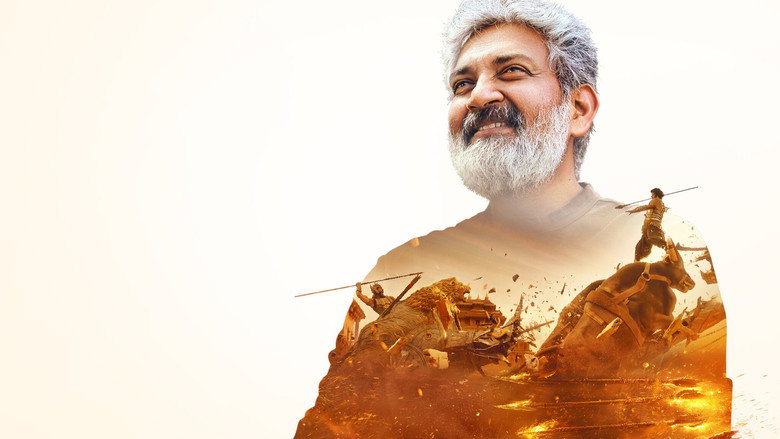 A behind-the-scenes shot of Rajamouli directing an action sequence, his arms outstretched as he demonstrates a movement to his actors