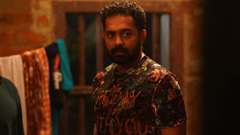 A tense standoff in a dimly lit warehouse, the camera angled low to emphasize the looming figures of Asif Ali and Vinayakan.