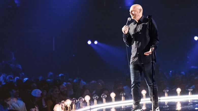 A dimly lit stage with a microphone stand at center, the audience a blur of faces in the dark.