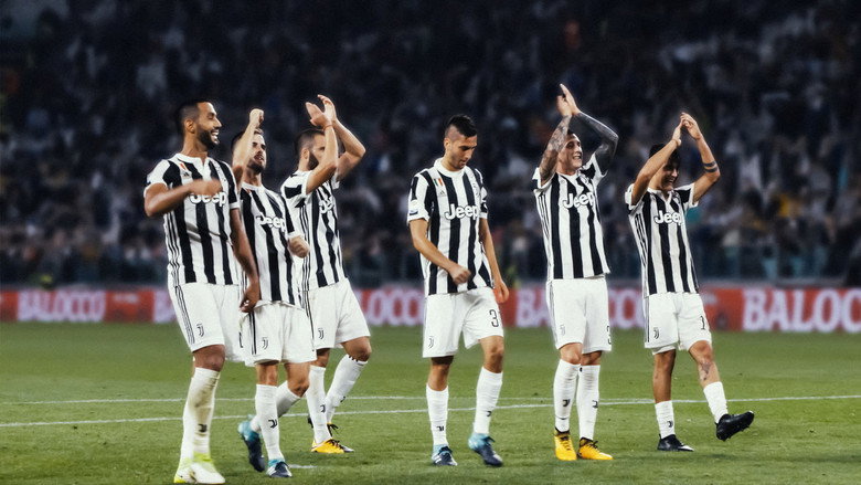 A Juventus player in mid-stride during training, his face obscured by the low angle of the camera, the black and white stripes of his kit stark against the green of the pitch.