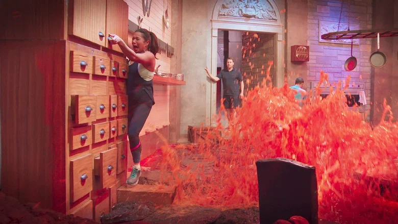 Contestants mid-air, arms outstretched, as they attempt to cross a room without touching the glowing orange "lava" below