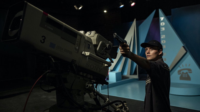 A young man in a TV studio, his face half-lit by the glow of monitors, gripping a gun with trembling hands