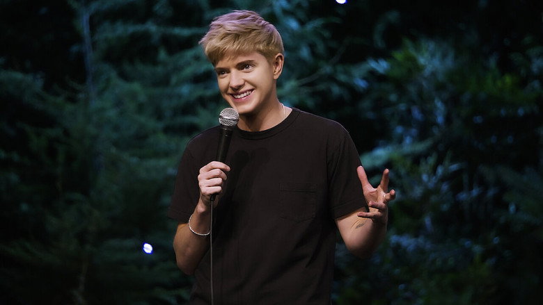 Mae Martin stands on stage at the Vogue Theatre, surrounded by lush greenery that evokes a forest setting, their expression caught between a joke and a revelation.