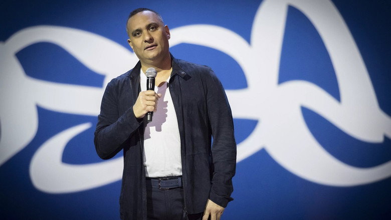 Russell Peters on stage, mid-performance, arms outstretched as he mimics an accent