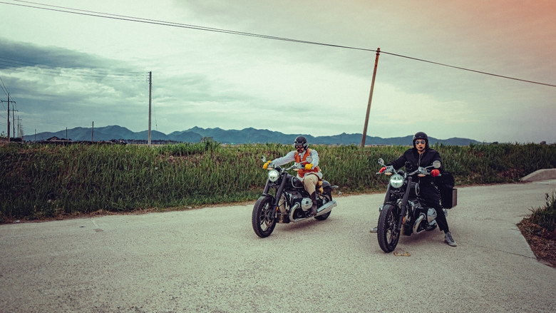 Two men on motorcycles pause at a scenic overlook, the Korean countryside stretching out behind them in soft focus