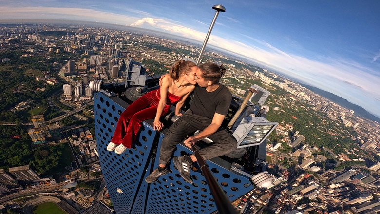 Angela and Ivan perched on a narrow ledge, the city sprawling beneath them like a circuit board.