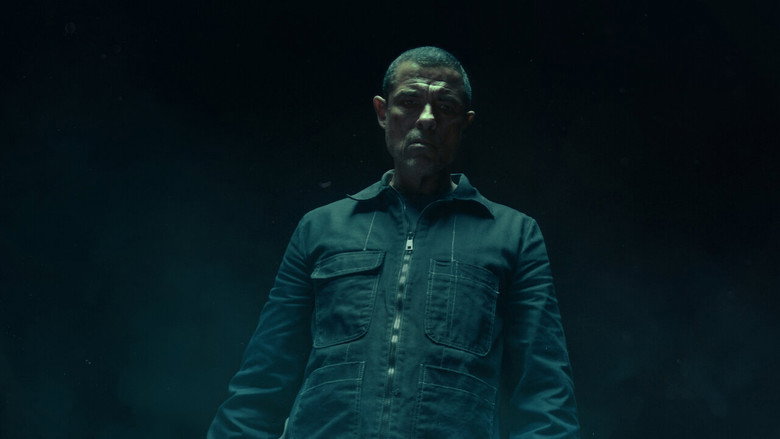A man in a dark coat stands in a snowy forest, his breath visible in the cold air, his face half-lit by an unseen light source.
