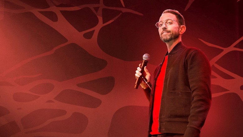 Neal Brennan on stage, bathed in green light, his face half in shadow as he delivers a punchline