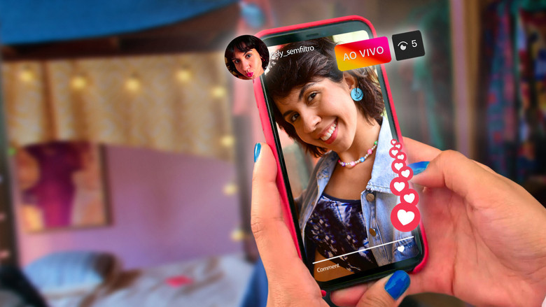 A close-up of Marcely’s phone screen, her face reflected in the glass as she records a video, the soft glow of the ring light casting her in an unnatural hue