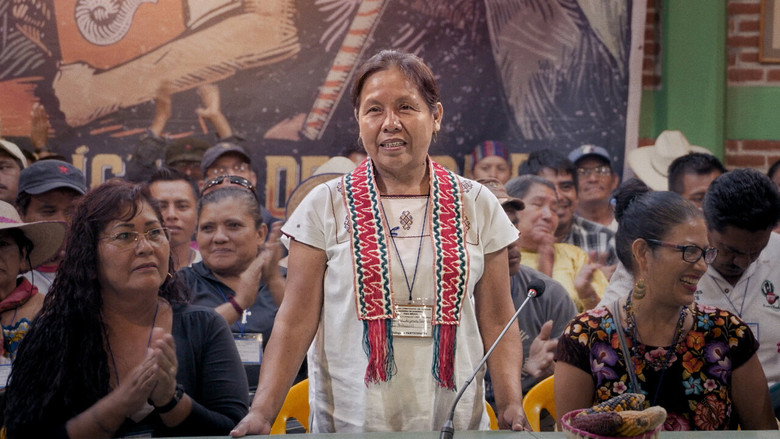 A group of Indigenous women in traditional dress stand together, their faces turned toward the camera with quiet determination.