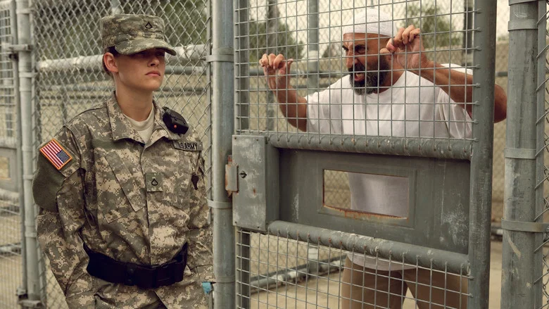 A guard in uniform stands in a dimly lit cell block, her face half in shadow.