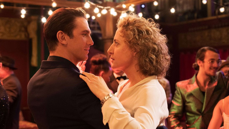 A dimly lit room where Alma and Tom sit across from each other at a table, the glow of a lamp casting long shadows that make their faces half-hidden, half-revealed.