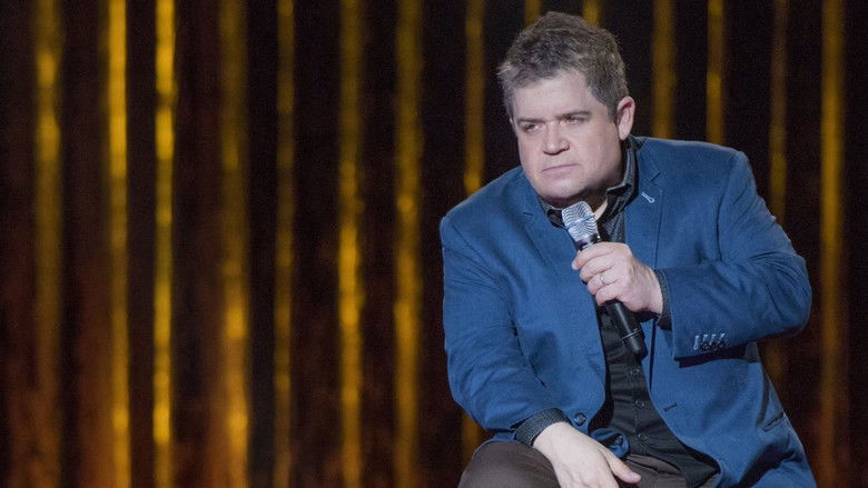 Patton Oswalt on stage, mid-gesture, bathed in warm light, the audience a blur of faces beyond the spotlight