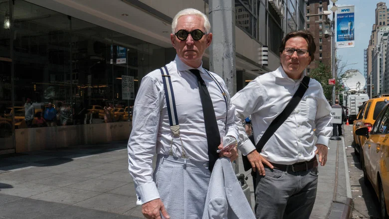 Roger Stone, in a pinstripe suit and American flag tie, smirks at the camera while seated in a dimly lit room, the glow of a lamp casting long shadows.