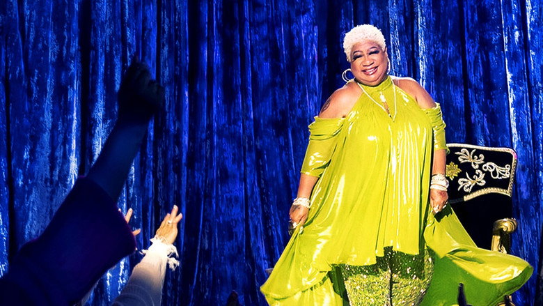 Luenell on stage, mid-laugh, her arms outstretched as if inviting the audience into her world