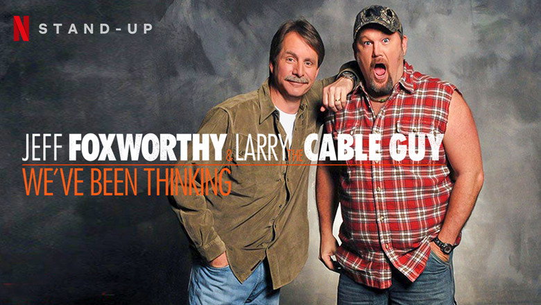Two men on stage, one in a red shirt gesturing with a microphone, the other leaning on a stool, both mid-laugh