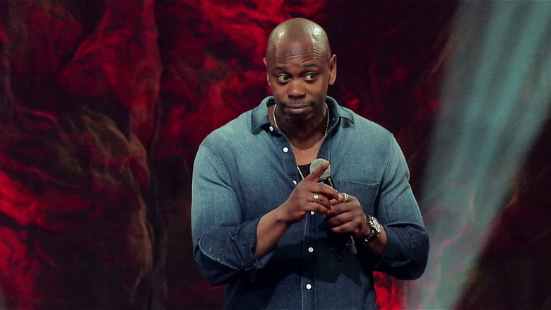 Chappelle on stage, bathed in blue light, mid-gesture