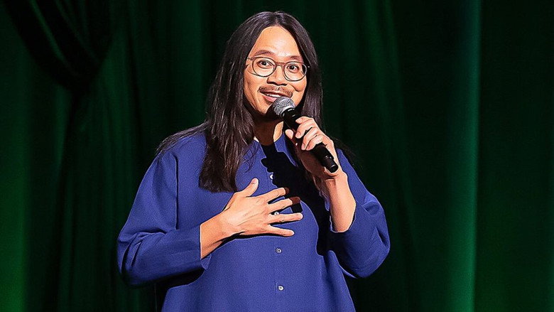 Sheng Wang on stage, mid-joke, his hands gesturing toward an invisible pint of berries