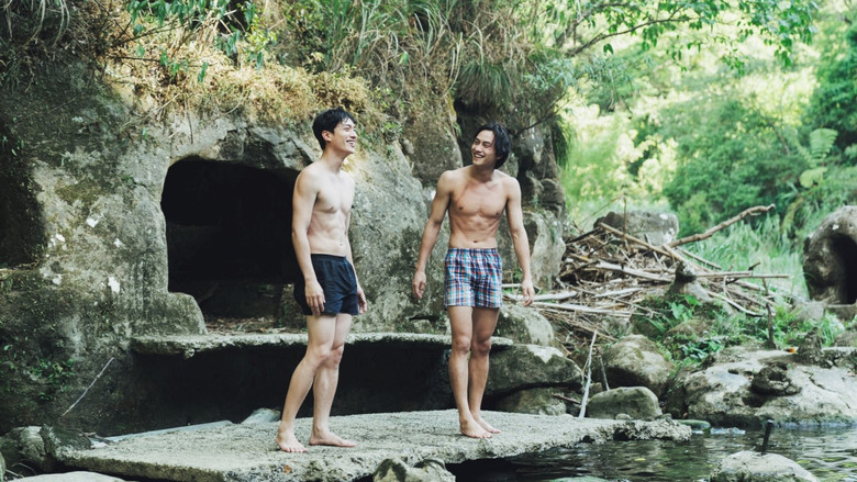 Two men sit on a rocky shore at dusk, the waves crashing behind them, their faces half-lit by the fading sun.
