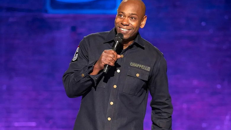 Chappelle on stage at the Lincoln Theatre, bathed in warm light, his expression caught between a smirk and something more contemplative