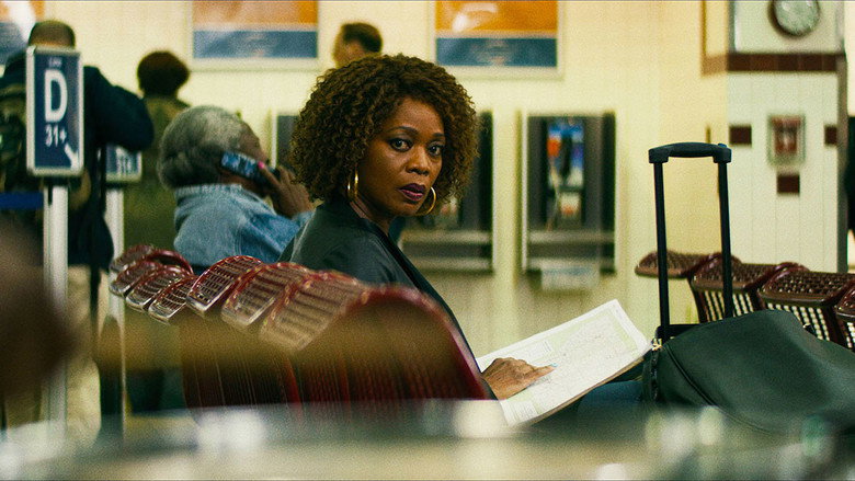 A woman in a red coat stands alone at a bus station, her back to the camera, as if poised between two versions of her life.