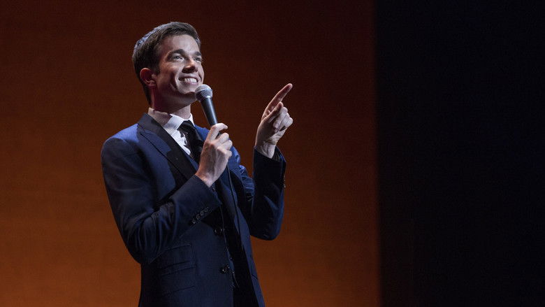 Mulaney mid-performance, bathed in warm stage light, his expression caught between a grin and a grimace