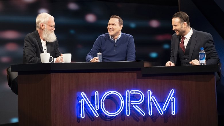 Norm Macdonald and Adam Eget sit across from a guest in a dimly lit, wood-paneled room, the camera capturing the quiet awkwardness of the moment.