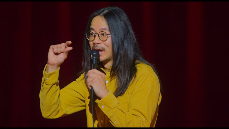 Sheng Wang on stage, bathed in soft blue light, mid-monologue about the absurdity of modern life