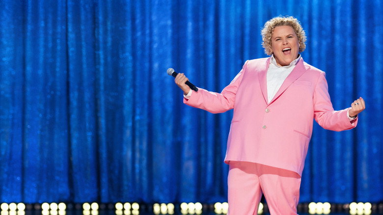 Fortune Feimster on stage in a pink suit, mid-laugh, arms outstretched