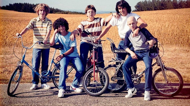 A group of five boys sitting on a hillside at dusk, their faces half-lit by the fading sun, sharing a quiet moment.