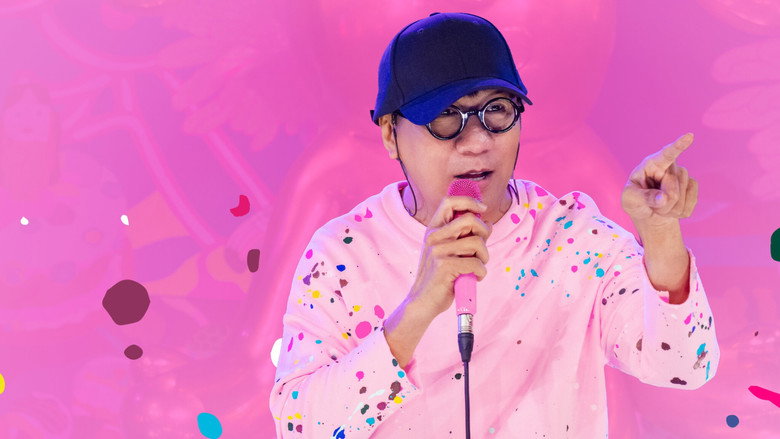 Udom Taephanich mid-gesture, spotlight catching the sweat on his forehead, the audience a blur of laughter