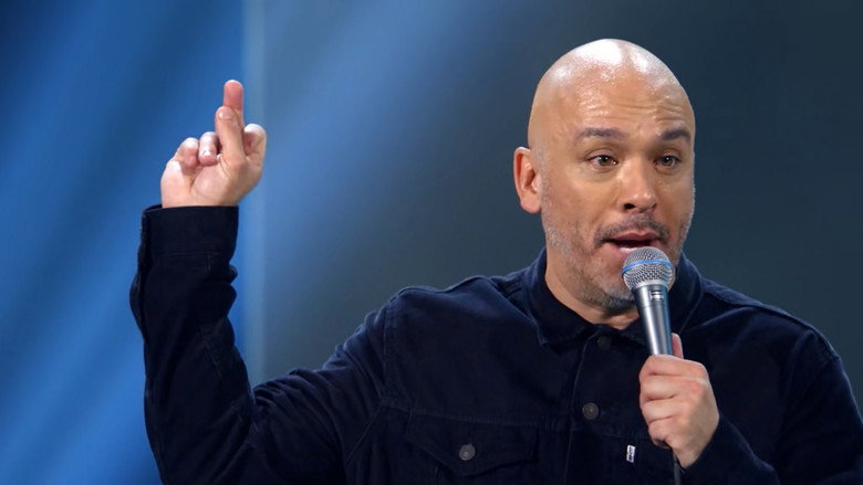Jo Koy on stage, arms outstretched, bathed in warm light, the arena audience a blur of faces behind him.