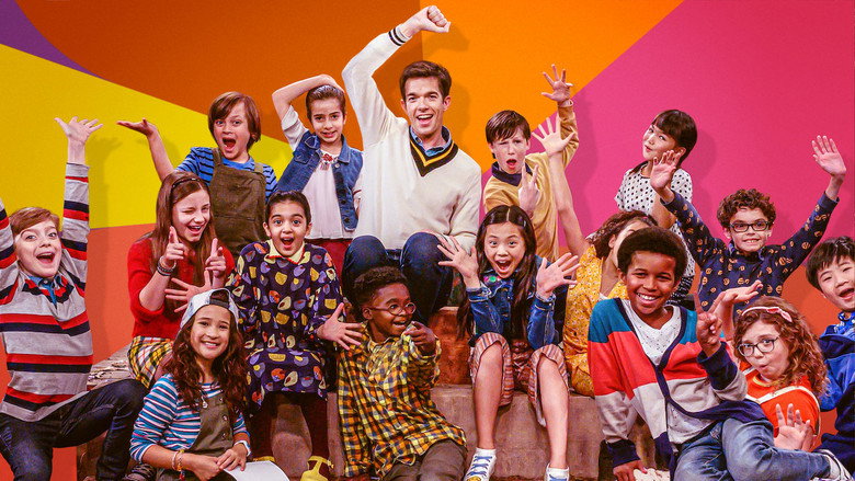 A group of kids in colorful outfits sit in a semicircle, grinning at the camera, while John Mulaney stands behind them in a suit, looking delighted and slightly out of place.