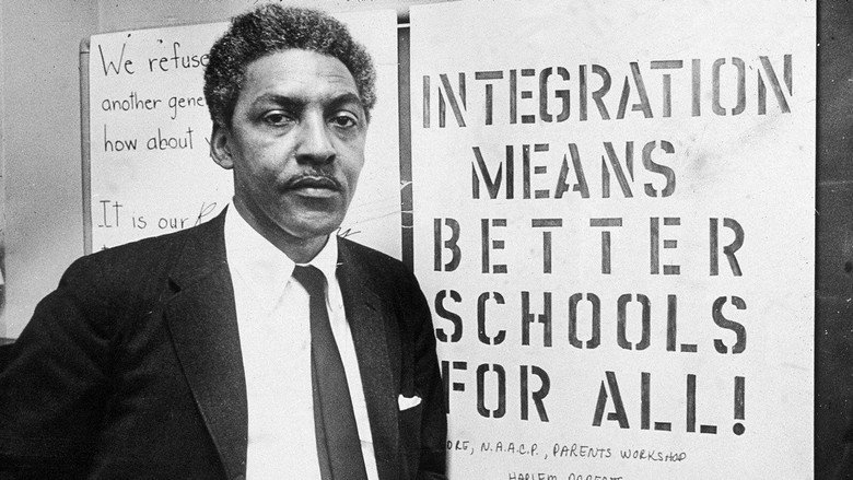 A black-and-white archival image of Bayard Rustin speaking at a podium, his hands raised in emphasis, surrounded by microphones.