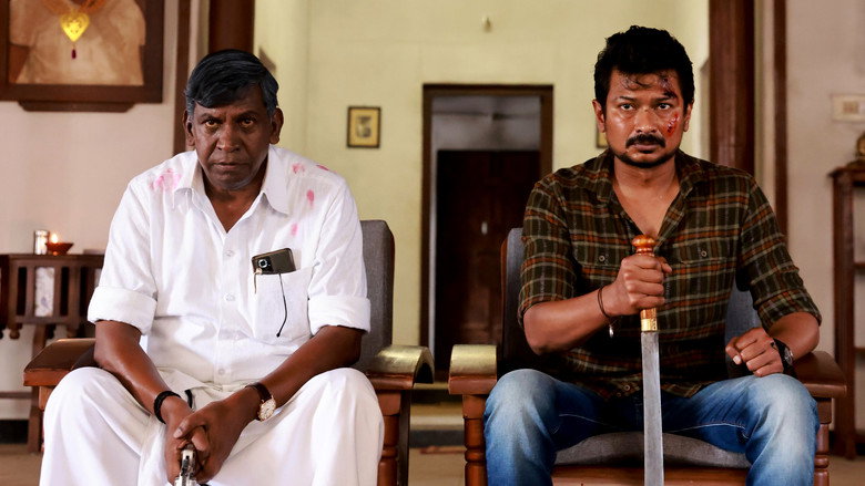 A tense standoff in a dimly lit room, where Vadivelu’s character stands defiantly over a wooden chair, his shadow stretching long across the floor.