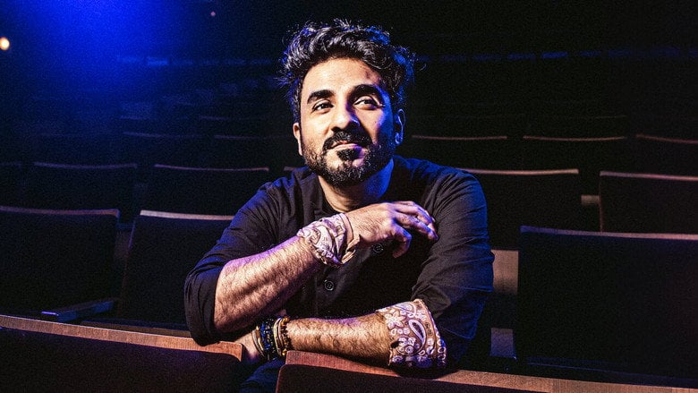 Vir Das on stage, mid-gesture, bathed in warm light, the audience a blur of faces beyond the spotlight