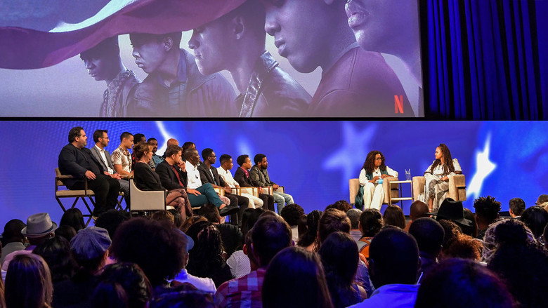 A dimly lit roundtable discussion, the exonerated men and their actors seated together, their expressions a mix of solemnity and quiet resolve.