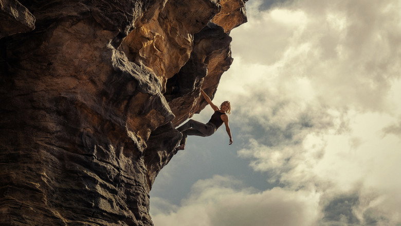 Charlize Theron climbing a jagged rock face, her face streaked with dirt and sweat, the Australian wilderness stretching out behind her.