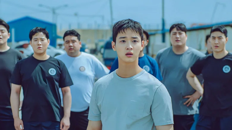 A quiet moment in the wrestling gym, where Baek-doo sits alone on the sand, his back to the camera, the warm light filtering through the windows.