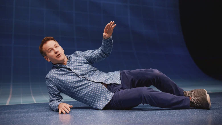 Birbiglia on stage, the graph-paper backdrop lit to resemble rippling water