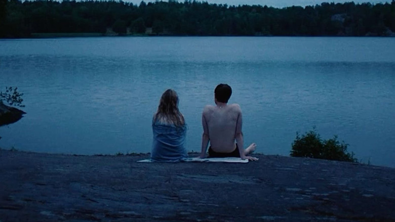 Agnes and Noel sit on a rooftop at dusk, their shoulders barely touching, the city sprawling beneath them like a living thing.