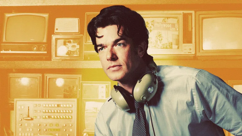 A wood-paneled set bathed in warm, earthy tones, with John Mulaney standing at a desk, mid-monologue, while Richard Kind lurks in the background, holding a stack of index cards.