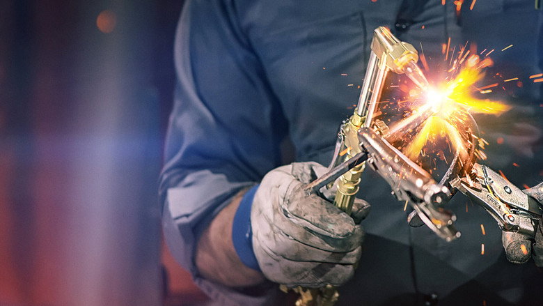A welder’s torch casts a blue-white glow, sparks cascading like a tiny meteor shower against the dark backdrop of the workshop