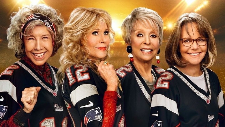 Four older women in colorful winter coats, laughing and huddled together in a stadium concourse.
