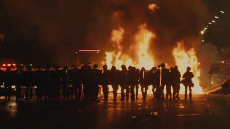 A sea of protesters fills a city square, their flags and banners a blur of green, yellow, and red under a hazy sky.