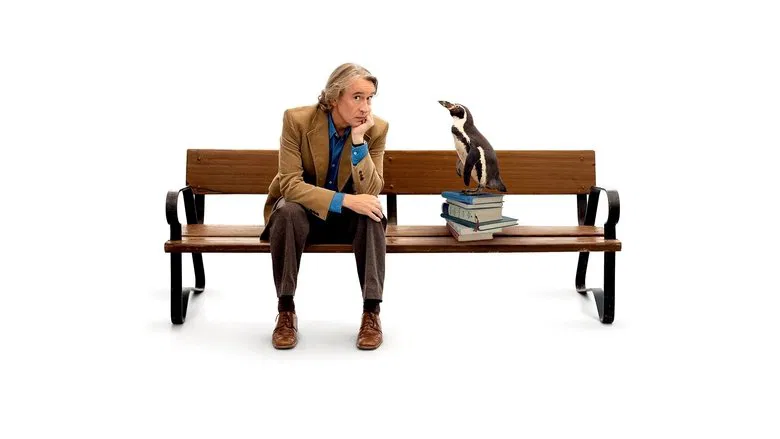 Steve Coogan, in a rumpled sweater, kneels in a dimly lit classroom, holding a small fish out to a penguin perched on a desk. The bird tilts its head, considering the offering, while a group of students watches in quiet awe.
