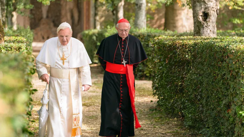 Two men in papal vestments walk side by side through the Vatican gardens, the sunlight casting long shadows.
