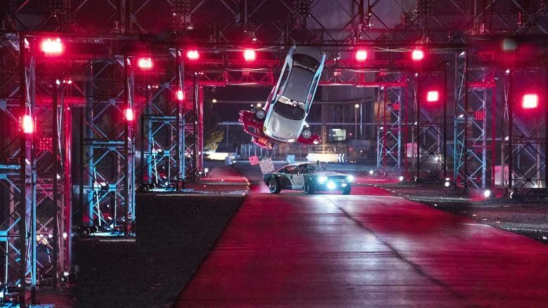 A custom car mid-air, soaring over a massive dirt ramp, its wheels spinning freely against the sky