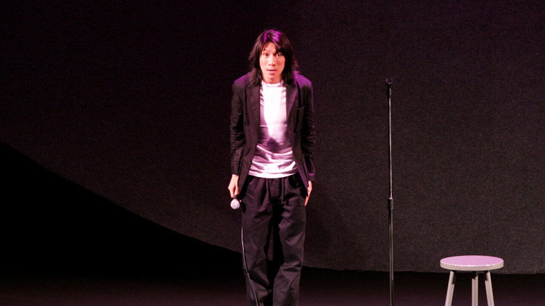 A dimly lit stage with a single microphone stand, the audience blurred in the foreground